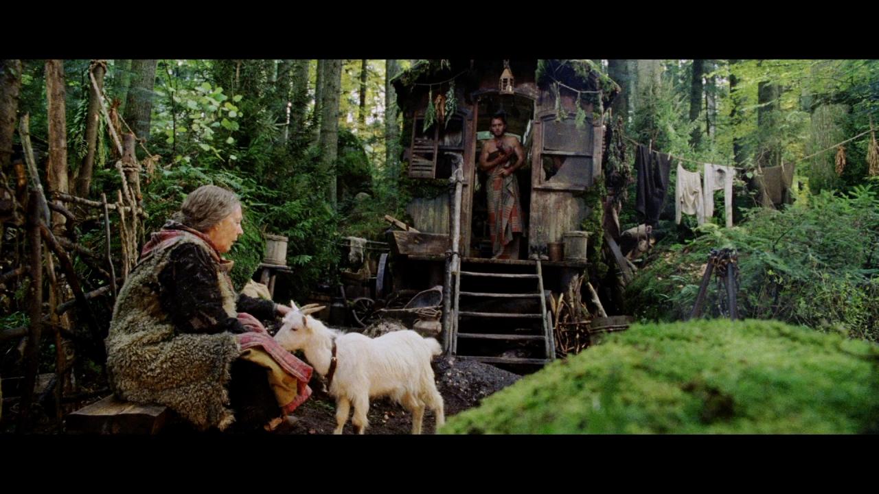 Eine alte Frau kümmert sich um eine Ziege. Im Hintergrund tritt ein junger Mann spärlich bekleidet aus einer markant gestalteten Hütte.